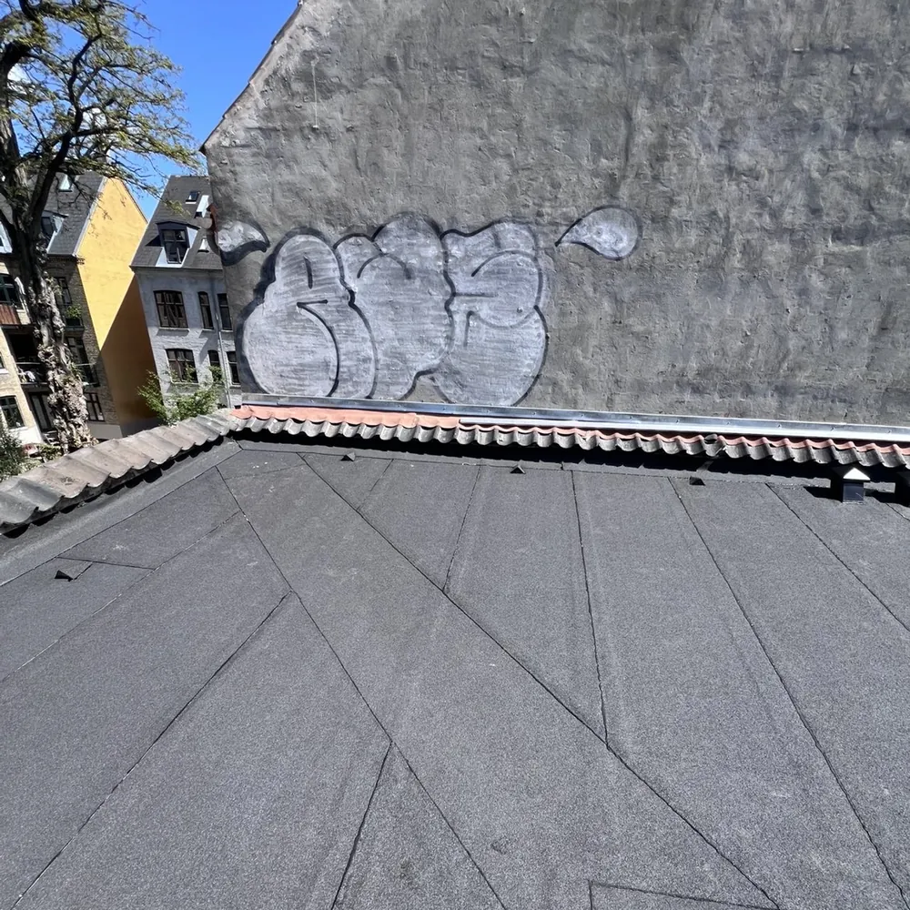Samme tagflade med nyt tagpap i én flade og nye udluftningshætter, set mod den samme tegltop og den samme gavl med graffiti